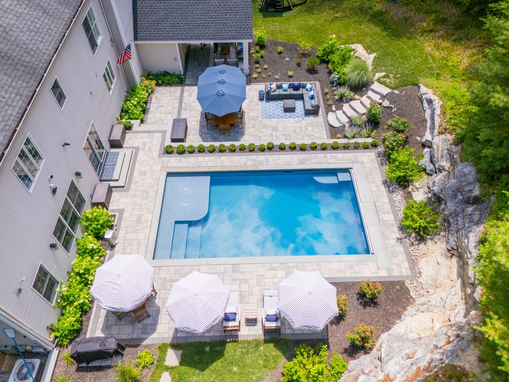 Overhead view of a paver pool patio with striped umbrellas, lounge seating, and a ledge rock border