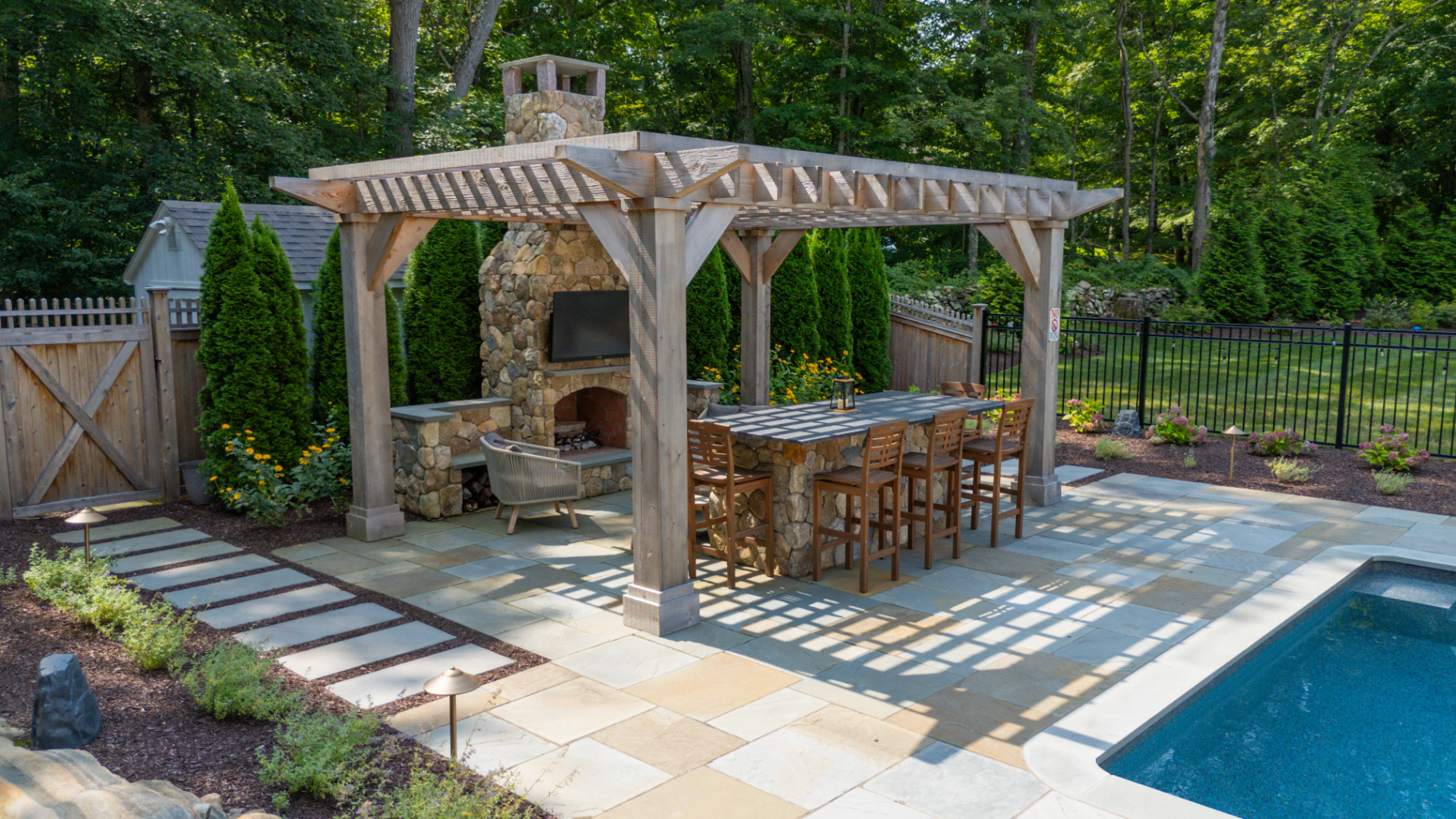 An outdoor living destination with timber pergola, stone fireplace, bar seating, and pool.